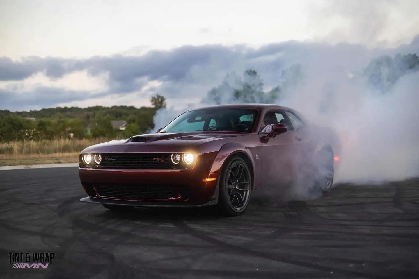 Dark red Dodge Challenger with its headlights on, surrounded by tire smoke on a paved lot