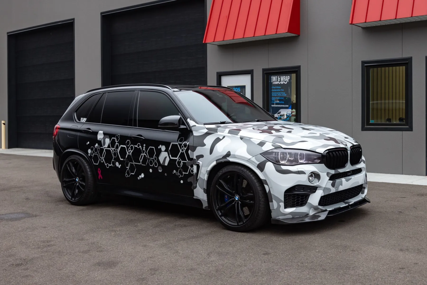 BMW X5 SUV with a white, gray and black camouflage-pattern wrap parked in front of a gray building with red awnings