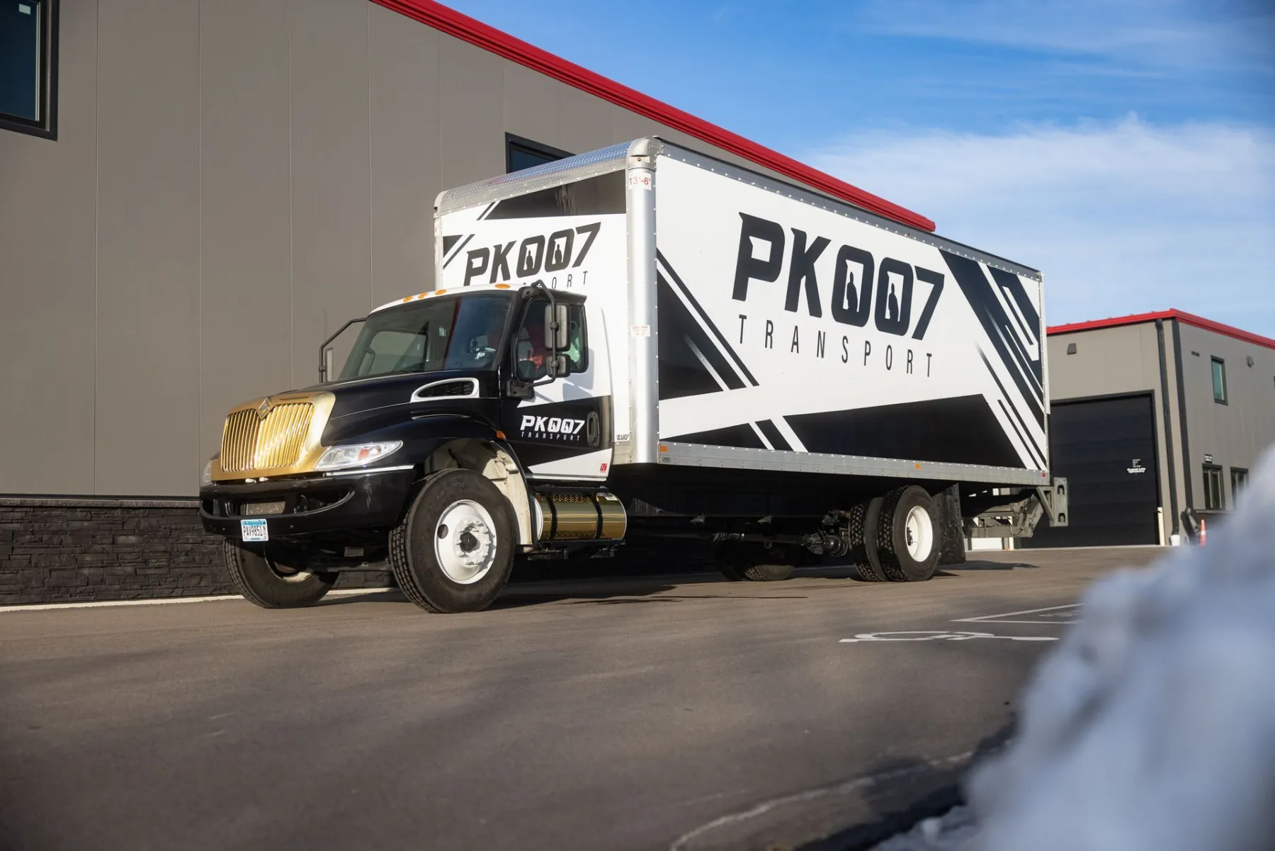 Low-angle view of a box truck with black and white 'PK007 Transport' graphics parked in front of a gray building with red trim