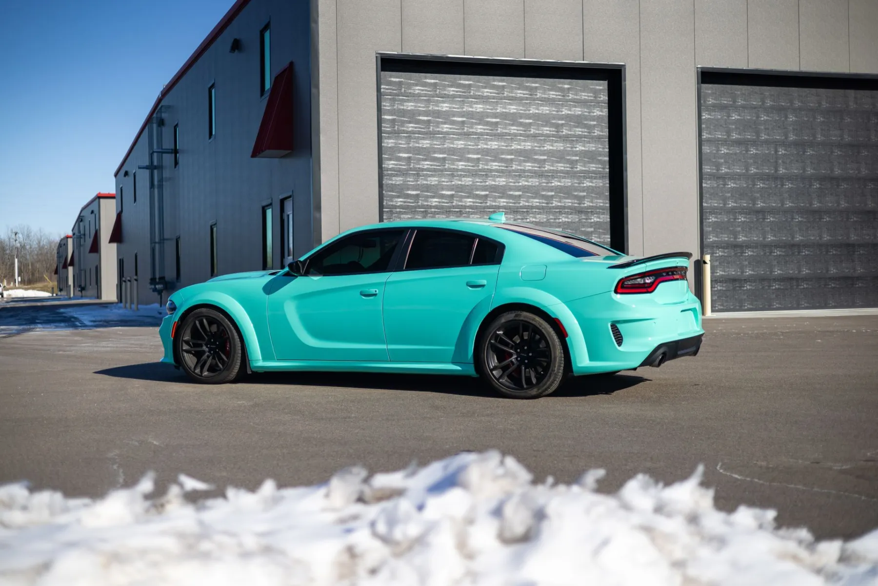 Teal Dodge Charger parked in front of a gray building with bay doors, a snow bank in the foreground