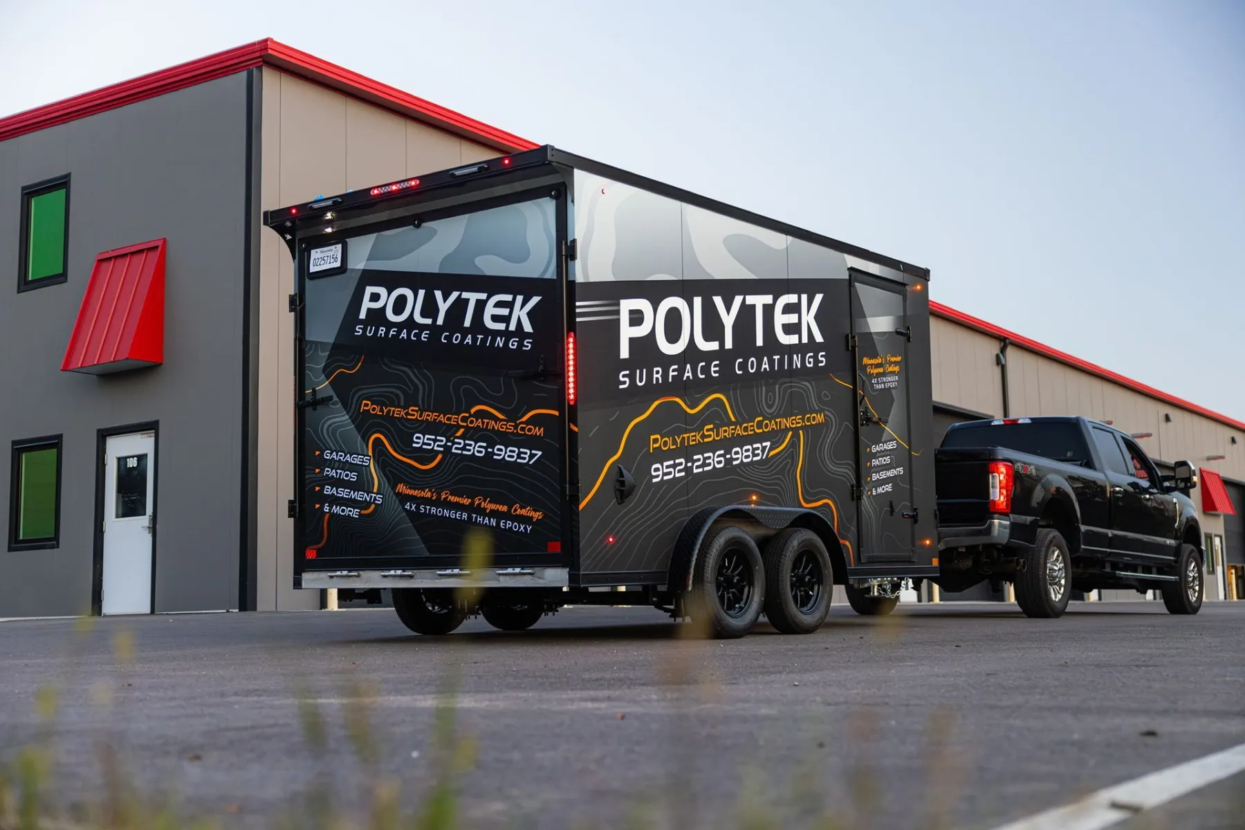 Enclosed trailer wrapped in black 'Polytek Surface Coatings' graphics hitched to a black pickup, parked in front of a gray building with red awnings