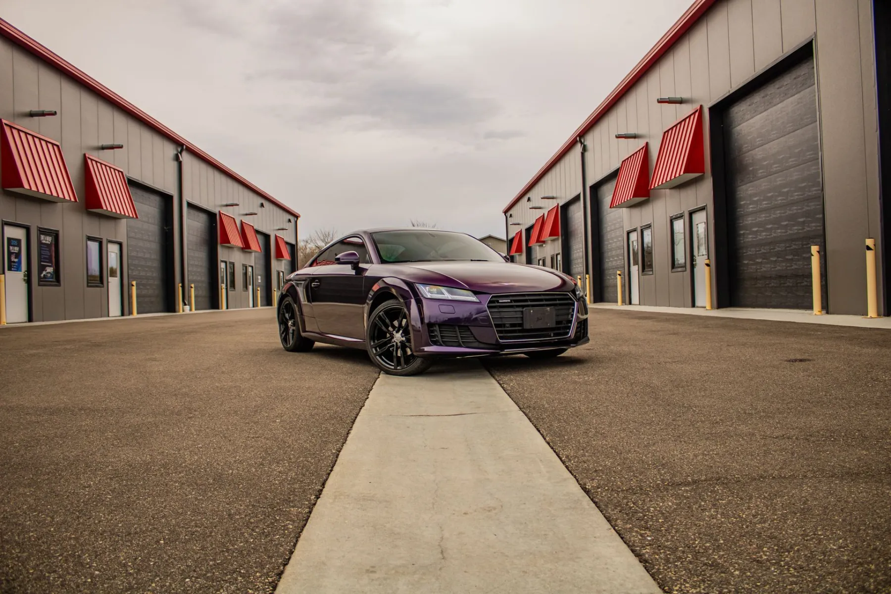 Purple Audi TT parked on a concrete strip between two rows of gray buildings with red awnings and bay doors