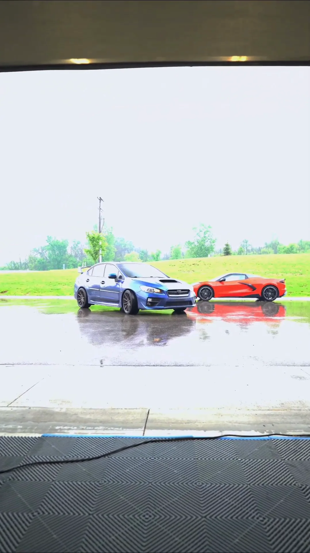 Blue Subaru WRX and a red sports car outside the open bay door in the rain