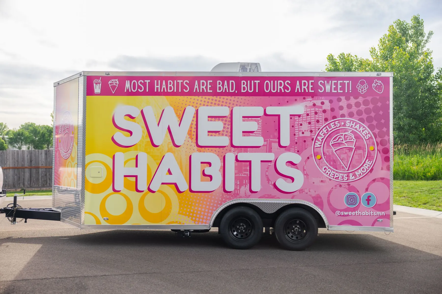 Enclosed trailer wrapped in pink and yellow 'Sweet Habits' graphics parked on pavement with trees behind