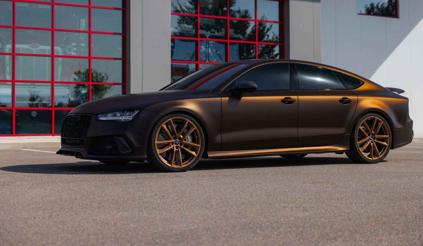 Audi RS7 with a matte brown finish and bronze wheels parked in front of a gray building with red-framed windows